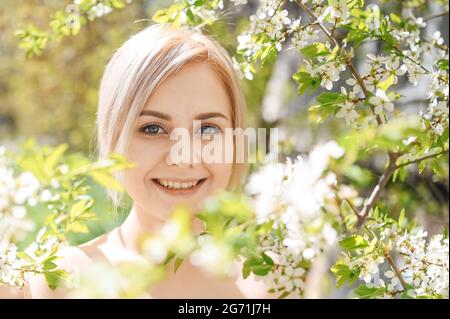 Jeune belle heureuse femme blonde souriante posant dans le jardin de printemps fleuri avec des fleurs blanches. Pommier ou cerisier en fleurs. Concept de cosmétiques naturels, jeunes, peau et dents saines Banque D'Images