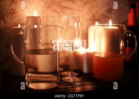 Pichets en verre avec boissons et gobelets vides placés près des bougies allumées sur la table dans la pièce sombre la nuit Banque D'Images