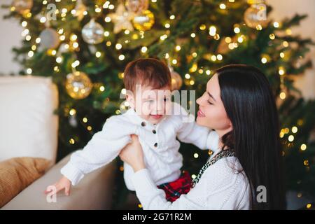 Jeune femme souriant et jouant avec un garçon mignon sur fond flou de conifères décorés le jour de Noël à la maison Banque D'Images