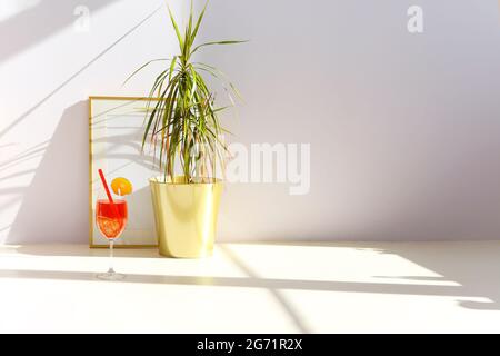 Verre de cocktail de fruits frais placé sur le sol près de la plante en pot et cadre vide contre le mur blanc dans la salle ensoleillée Banque D'Images