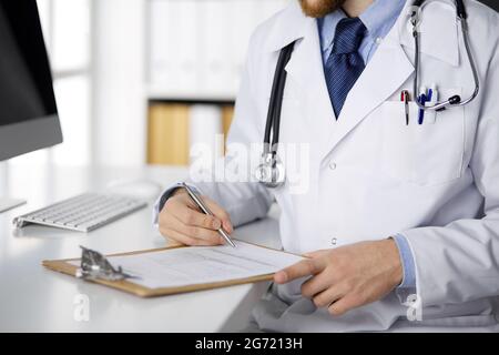 Médecin sympathique à barbe rouge assis et écrivant avec une planchette à pince en clinique, en gros plan. Concept de médecine Banque D'Images