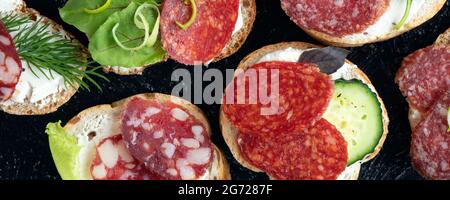 banderole avec des sandwiches ou des en-cas de type gros plan avec différents types de saucisses et de légumes sur un fond noir de texture. Mise au point douce. Vue de dessus. pose à plat Banque D'Images