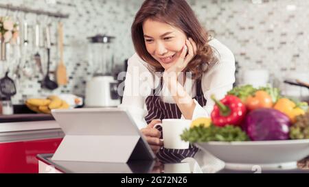 Femme asiatique mignonne d'âge moyen tenant une tasse de café et utilisant un ordinateur de tablette se connecter à Internet dans la cuisine avec un visage souriant et de manière heureuse. Concept Banque D'Images
