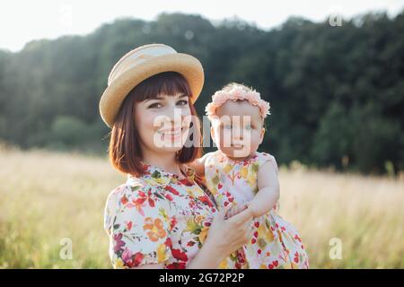 Bonne mère et petite fille, portant des robes colorées similaires, ayant du plaisir dans le champ d'été au coucher du soleil doré. Maman et enfant souriant sur les mains, regardant l'appareil photo. Campagne, bonne enfance Banque D'Images