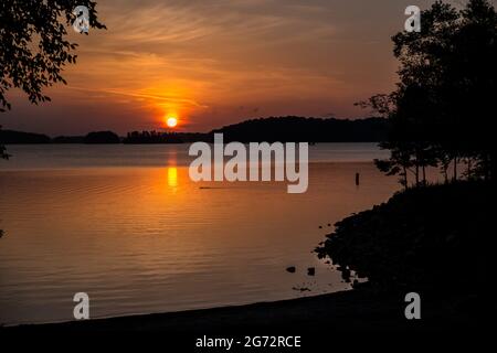 Un lever de soleil orange profond au-dessus du lac avec les îles et le rivage avec des arbres silhouettés et une famille de canards nageant en été sur le lac Lani Banque D'Images