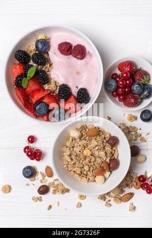 Vue de dessus de bols avec flocons d'avoine, framboise, fraise, myrtille, groseille, yaourt maison aux amandes, noisettes et fruits roses sur un filet de bois blanc Banque D'Images