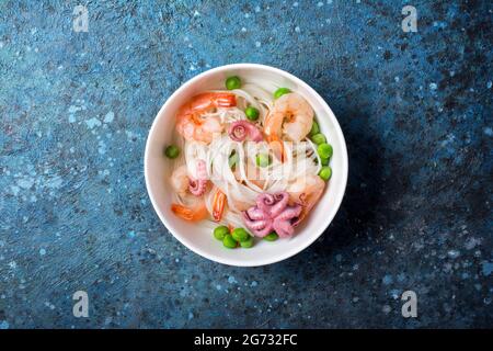 Vue de dessus du bol avec nouilles de riz chinoises, crevettes savoureuses, bbe octopus et pois verts sur fond de béton bleu Banque D'Images