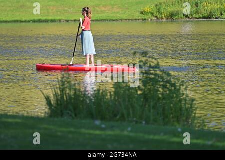 Une jeune femme traverse le lac olympique dans le parc olympique de Munich le 10 juillet 2021. Stand Up Paddling.SUP Stand Up Paddling (SUP), également appelé stand up Paddling ou stand up paddle surf, est un sport aquatique dans lequel un athlète se tient debout sur une sorte de planche de surf et des pagaies avec une paddle. Banque D'Images