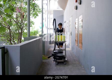 Une femme de ménage asiatique en uniforme bleu pousse le chariot de ménage dans le couloir devant la chambre d'hôtel. Le panier à linge, le nettoyant pour vitres, les gants en caoutchouc, Banque D'Images