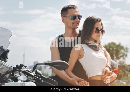 Joyeux couple souriant amoureux des lunettes de soleil. Beau homme et jolie femme sont embrassant. Les jeunes gens attrayants posent sur la moto. Horizon Banque D'Images