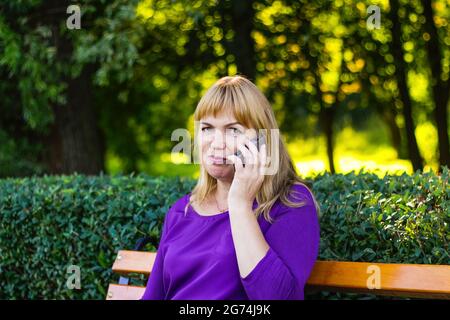 Flou artistique Femme blonde caucasienne parlant, parlant au téléphone à l'extérieur, à l'extérieur. femme de 40 ans en chemisier violet dans le parc sur banc. Adulte usi Banque D'Images