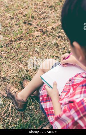 Vue arrière. Enfant utilisez un stylo pour écrire sur un carnet en bois dans le parc. À l'extérieur, en journée, en plein soleil. Utilisez-le pour l'apprentissage ou l'éducation Banque D'Images