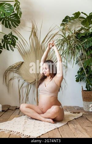 Belle femme enceinte en maillot de bain sport beige faisant du yoga à la maison Banque D'Images