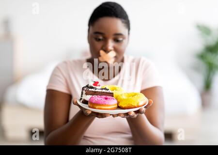 Restrictions alimentaires. Femme noire curvy avec bandage adhésif sur sa bouche regardant la plaque de bonbons dans la faim à la maison Banque D'Images