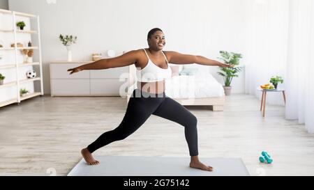 Yoga de force pour la perte de poids. Femme noire curvy debout dans l'asana guerrier, s'exerçant sur tapis de sport à la maison, panorama Banque D'Images