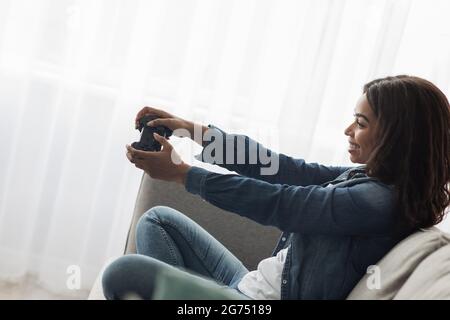 Passe-temps du week-end. Joyeuse afro-américaine dame jouant à des jeux vidéo en ligne, tenant le joystick, assis sur un canapé, vue latérale Banque D'Images