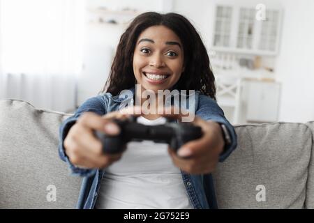 Week-end loisirs. Une femme afro-américaine surjoyée utilisant le joystick et jouant aux jeux vidéo, assise sur un canapé à la maison Banque D'Images