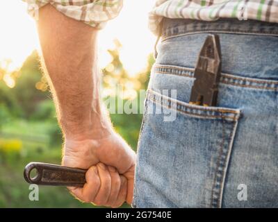 Beau homme en vêtements de travail, tenant des outils dans ses mains contre le fond des arbres, le ciel bleu et le coucher du soleil. Vue de l'arrière. Le travail et les employés Banque D'Images