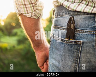 Beau homme en vêtements de travail, tenant des outils dans ses mains contre le fond des arbres, le ciel bleu et le coucher du soleil. Vue de l'arrière. Le travail et les employés Banque D'Images