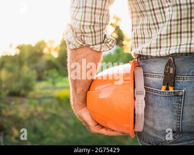 Homme attrayant dans des vêtements de travail, tenant des outils dans ses mains contre le fond des arbres, le ciel bleu et le coucher du soleil. Vue de l'arrière. Main-d'œuvre et emploi Banque D'Images