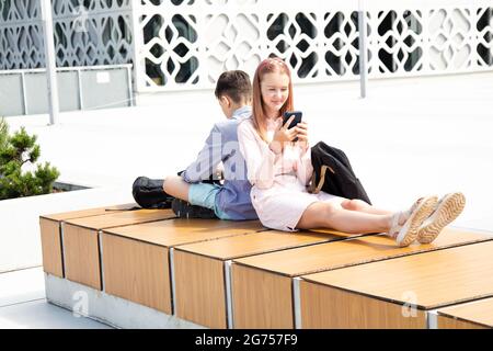 Filles et garçons écoliers assis avec des sacs à dos d'école sur un banc en bois, entre les murs en béton dos à dos, utiliser des appareils mobiles Banque D'Images