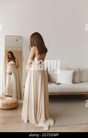 Une jeune femme enceinte belle dans une longue robe tendance est debout devant le miroir et regarde son ventre. Derniers mois de grossesse Banque D'Images