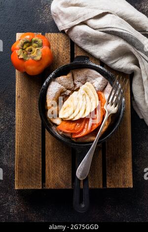 Petite crêpe hollandaise avec du persimmon frais et de la banane avec de la poudre de sucre glace dans une casserole rouge sur fond blanc de cuisine. Vue de dessus Banque D'Images