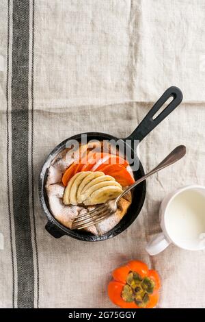 Petite crêpe hollandaise avec du persimmon frais et de la banane avec de la poudre de sucre glace dans une casserole rouge sur fond blanc de cuisine. Vue de dessus Banque D'Images