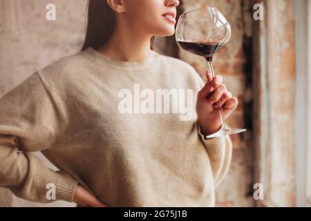 Crop Happy female en chandail sentant le vin rouge dans un grand verre sur fond gris Banque D'Images