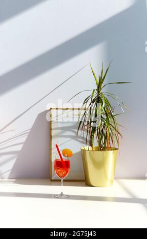 Un verre de cocktail de spritz d'Apperol est placé sur le sol près de la plante en pot et vide le cadre contre le mur blanc dans la salle ensoleillée Banque D'Images