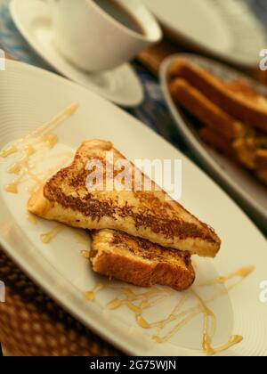 Deux toasts français servis avec du miel sur une assiette blanche pour le petit-déjeuner ou le brunch, en arrière-plan une tasse de café noir et une pile de toasts français. Banque D'Images