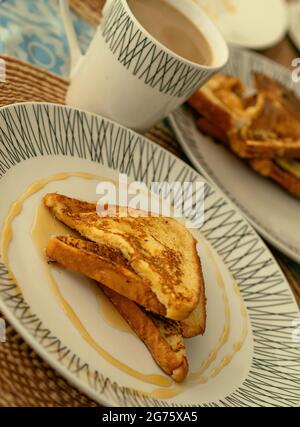 Deux toasts français servis avec du miel sur une assiette blanche pour le petit-déjeuner ou le brunch, en arrière-plan une tasse de latte de café et une pile de toasts français. Banque D'Images