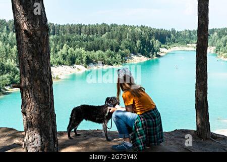 Vue arrière de la jeune femme blonde voyageur en chapeau et gingembre t-shirt debout sur la falaise de bleu turquoise lac ou rivière et conifères avec duveteux d Banque D'Images