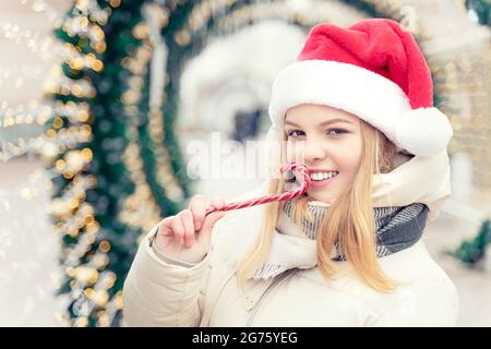 Portrait de Noël d'une adolescente blonde souriante portant un chapeau de père noël rouge tenant une canne à sucre à l'extérieur Banque D'Images