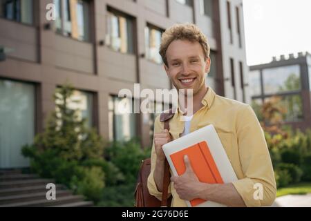 Beau étudiant souriant portant des vêtements décontractés tenant un ordinateur portable et livre regardant l'appareil photo, marchant à l'université. Retour à l'école, concept d'éducation Banque D'Images