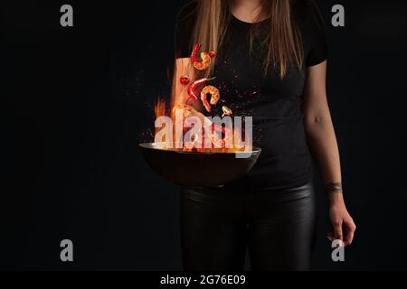 Queues de crevettes et légumes planant dans le feu au-dessus du wok dans les mains des femmes Banque D'Images