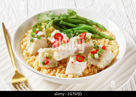 Bacalhau com todos, morue au bulgur, haricots verts cuits à la vapeur, saupoudrée de piments forts et d'oignons verts dans un bol blanc sur une table en bois, balkans Banque D'Images