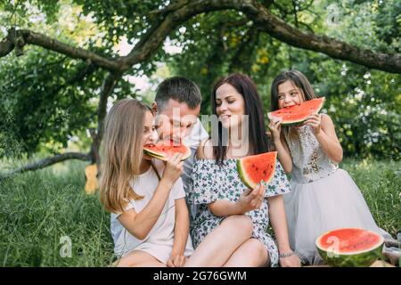 Parents joyeux avec des filles en costume d'été assis ensemble dans le jardin vert et appréciant la pastèque douce. Pique-nique à l'extérieur. Temps de relaxation. Banque D'Images