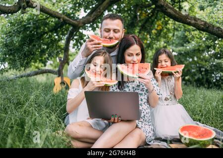 Famille caucasienne positive ayant pique-nique d'été manger du melon d'eau. Femme tenant un ordinateur portable sans fil pendant que deux filles et un mari l'embrassant. Concept de bonheur. Banque D'Images