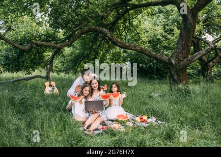 Jeunes parents avec deux petites filles ayant un appel vidéo sur ordinateur portable pendant le pique-nique dans le jardin verdoyant. Famille heureuse parlant avec des parents en ligne. Concept de personnes et de technologie. Banque D'Images