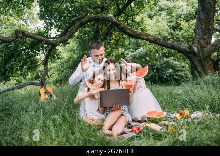 Jeunes parents avec deux petites filles ayant un appel vidéo sur ordinateur portable pendant le pique-nique dans le jardin verdoyant. Famille heureuse parlant avec des parents en ligne. Concept de personnes et de technologie. Banque D'Images