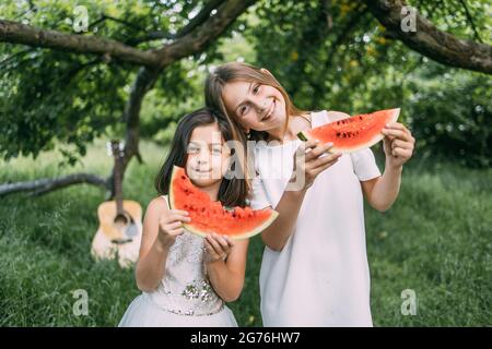 Jolis enfants en robe d'été appréciant la douce pastèque à l'extérieur. Deux filles debout dans un jardin verdoyant, regardant l'appareil photo et souriant sincèrement. Connexion entre sœurs. Concept de famille et d'amour. Banque D'Images