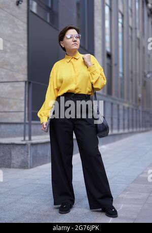 Femme d'affaires caucasienne brune dans un pantalon noir et un chemisier jaune debout devant le bâtiment de bureau. Contexte urbain, finance ou concept d'assurance. Photo verticale de haute qualité Banque D'Images