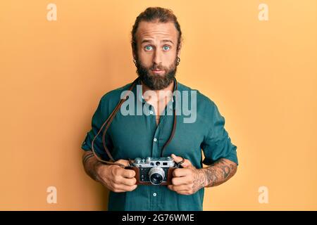 Beau homme avec la barbe et les cheveux longs tenant l'appareil photo vintage expression sans indice et confuse. Doute concept. Banque D'Images
