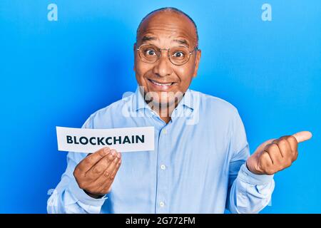 L'homme latin d'âge moyen tient le papier avec le message blockchain criant fier, célébrant la victoire et le succès très excité avec le bras relevé Banque D'Images