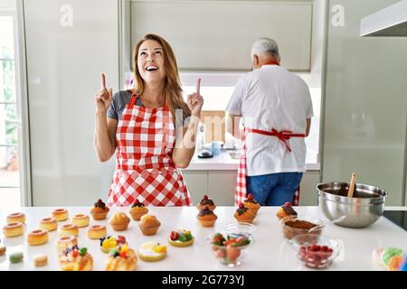 Femme hispanique d'âge moyen portant un tablier cuisant des pâtisseries maison stupéfait et surpris regardant vers le haut et pointant avec les doigts et les bras relevés. Banque D'Images