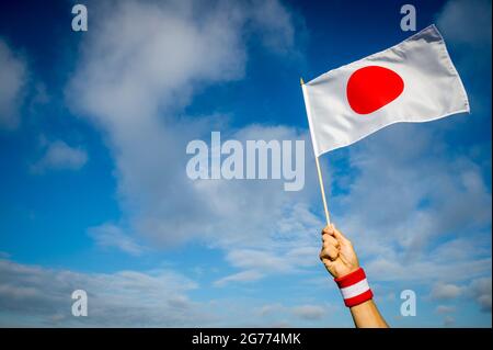 Main tenant le bracelet rouge et blanc japonais tenant le drapeau national du Japon en agitant dans un ciel bleu clair et ensoleillé Banque D'Images