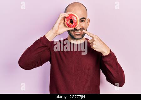 Jeune homme chauve tenant un savoureux donut coloré sur l'oeil souriant heureux pointant avec la main et le doigt Banque D'Images