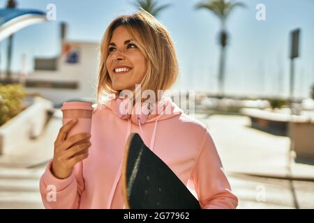 Jeune fille blonde patineuse souriante heureuse tenant skate boire un café à la ville. Banque D'Images