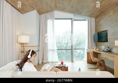 Jeune femme créative assise sur un canapé à la maison, écoutant de la musique et dessinant dans un carnet de croquis Banque D'Images